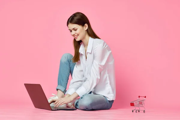 Mujer alegre con el ordenador portátil sentado en el suelo en el fondo rosa tecnología de Internet — Foto de Stock