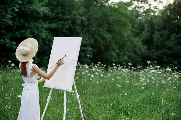 Μια γυναίκα σε ένα λευκό φόρεμα σε ένα χωράφι με λουλούδια ζωγραφίζει μια εικόνα — Φωτογραφία Αρχείου