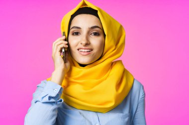 Sarı tesettürlü müslüman kadın telefonda pembe teknolojiyle konuşuyor.