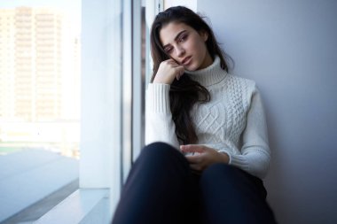 Pencerenin yanında beyaz kazaklı esmer.