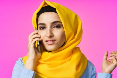 Sarı tesettürlü müslüman kadın telefonda pembe teknolojiyle konuşuyor.