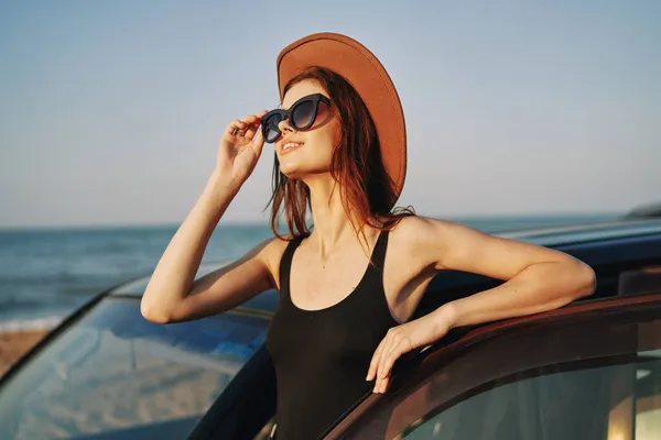 Fröhliche Frau mit Sonnenbrille und Hut auf dem Meer in der Nähe des Autos — Stockfoto