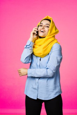 Sarı tesettürlü müslüman kadın telefon teknolojisi pembe arka planda konuşuyor.