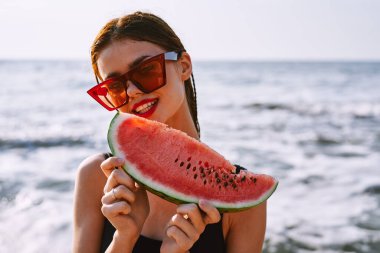 Güneş gözlüklü neşeli kadın okyanusta karpuz yiyor.