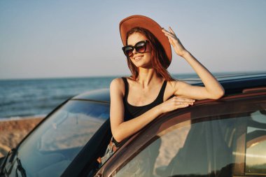 Güneş gözlüklü neşeli bir kadın ve arabanın yanındaki okyanusta bir şapka.