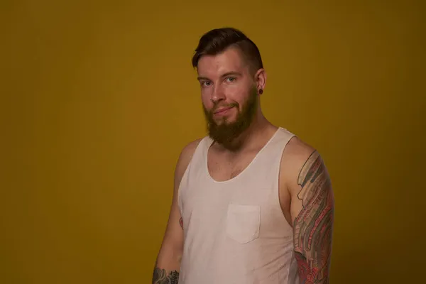 Brodaty mężczyzna w białej koszulce z tatuażami na ramionach, pozujący na poważny wygląd — Zdjęcie stockowe