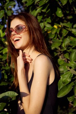 Siyah mayo giymiş neşeli kadın yeşil yapraklar doğada poz veriyor.