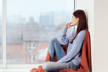 Hüzünlü yüzü olan kadın elinde ekoseyle pencerenin kenarında oturuyor.