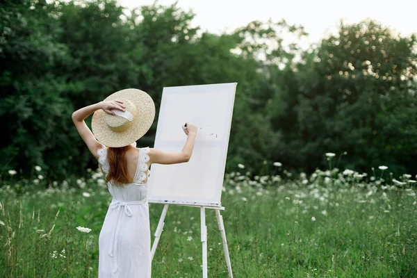 Una mujer en un vestido blanco en un campo con flores pinta un cuadro —  Fotos de Stock