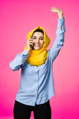 Sarı tesettürlü müslüman kadın telefon teknolojisi pembe arka planda konuşuyor.