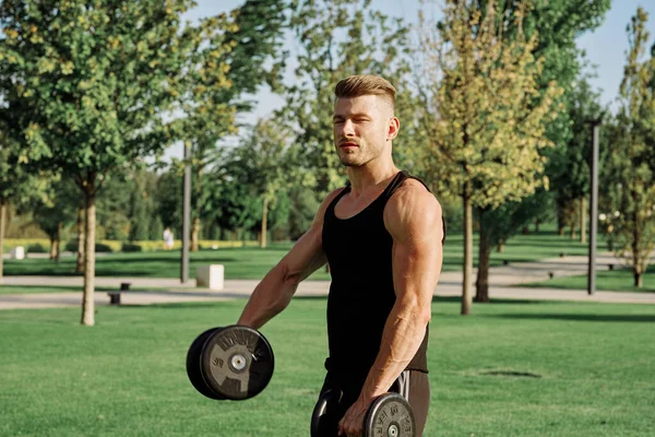 Atletyczny mężczyzna w czarnej koszulce z hantlami na treningu w parku — Zdjęcie stockowe