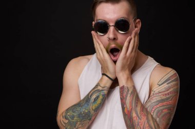 Kollarında dövme olan yakışıklı bir adam. Güneş gözlüğü takıyor. Geçmişi koyu renk.