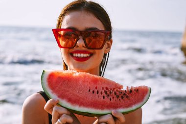 Güneş gözlüklü neşeli kadın okyanusta karpuz yiyor.