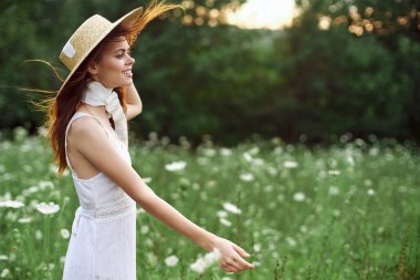 Çiçekli bir tarlada şapkalı güzel bir kadın yürüyor.