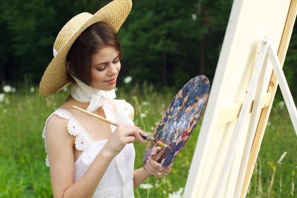 Hezká žena v klobouku umělec maluje obrázek na přírodu malovat — Stock fotografie