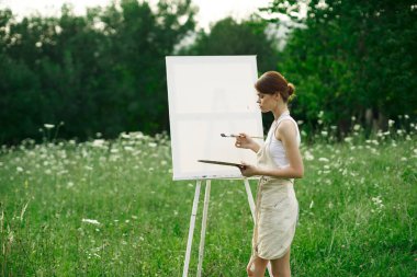 Kadın sanatçı, doğa manzarasında sehpada resim yapar.