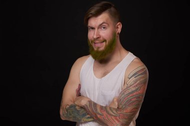 Kollarında dövmeleri olan beyaz tişörtlü şık sakallı bir adam.