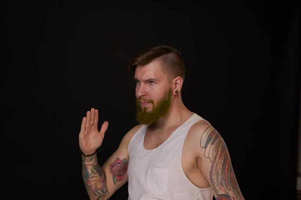 Homme barbu avec des tatouages sur les bras gesticulant avec ses mains fond sombre — Photo