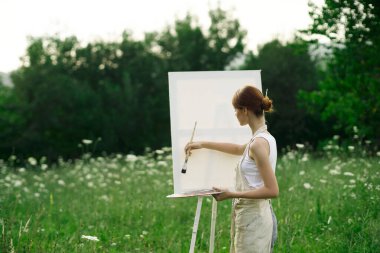 Kadın sanatçı, doğa manzarasında sehpada resim yapar.