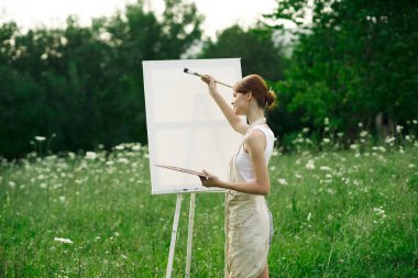 Kadın sanatçı, doğa manzarasında sehpada resim yapar.