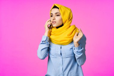 Sarı tesettürlü neşeli bir kadın. Elinde telefon, iletişim teknolojisi var.