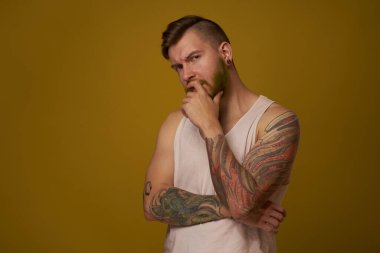 Kollarında dövmeleri olan beyaz tişörtlü sakallı adam ciddi görünüyor.