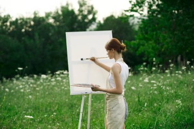 Kadın sanatçı, doğa manzarasında sehpada resim yapar.
