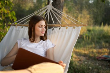 Neşeli kadın hamakta dizüstü bilgisayarlı internet seyahatiyle yatıyor.