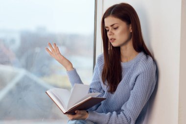 Kadın pencerenin yanında kitap okuyor, bir bardak içkiyle dinleniyor.