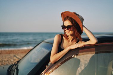Güneş gözlüklü neşeli bir kadın ve arabanın yanındaki okyanusta bir şapka.