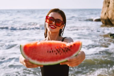Güneş gözlüklü neşeli kadın okyanusta karpuz yiyor.