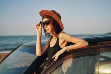 Güneş gözlüklü neşeli bir kadın ve arabanın yanındaki okyanusta bir şapka.