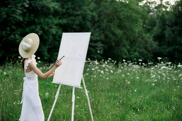 Una mujer en un vestido blanco en un campo con flores pinta un cuadro —  Fotos de Stock