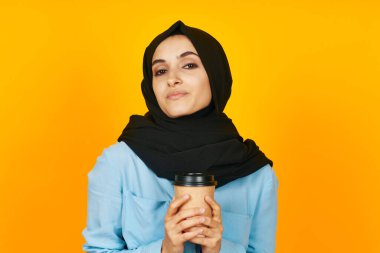 Neşeli kadın, elinde bir bardak kahve, yüzüne yakın, sarı arka plan.