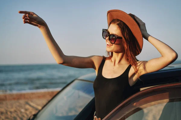 Χαρούμενη γυναίκα με γυαλιά ηλίου και ένα καπέλο στον ωκεανό κοντά στο αυτοκίνητο — Φωτογραφία Αρχείου