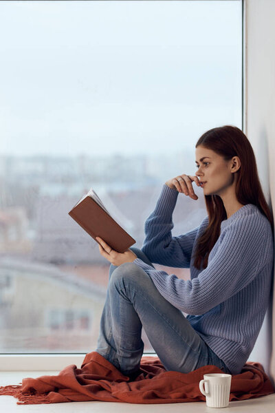 женщина читает книгу у окна с чашкой напитка отдыха