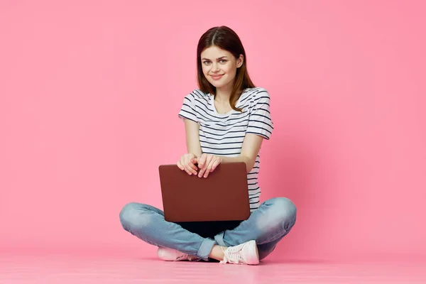 Mujer alegre con el ordenador portátil sentado en el suelo sobre un fondo rosa — Foto de Stock