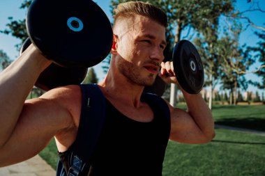 Parkda elinde dambıllar olan atletik bir adam yaşam tarzını eğitiyor.