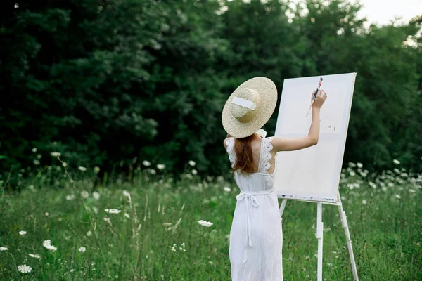 A woman in a white dress in a field with flowers paints a picture — Stock Photo, Image