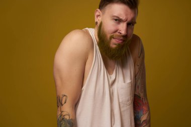 Beyaz tişörtlü, kollarında dövmeleri olan sakallı bir adam.