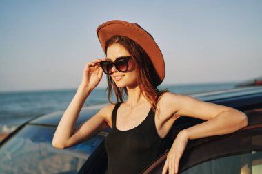 Güneş gözlüklü neşeli bir kadın ve arabanın yanındaki okyanusta bir şapka.