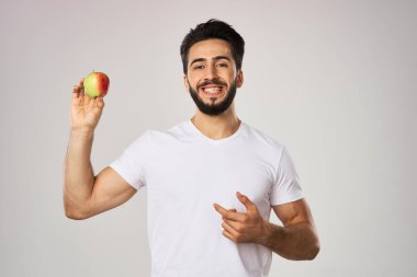 Beyaz tişörtlü, sağlıklı meyvelerin elinde elma olan neşeli bir adam.