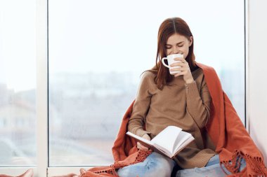 Pencere kenarında bir bardak içkiyle kitap okuyan bir kadın.