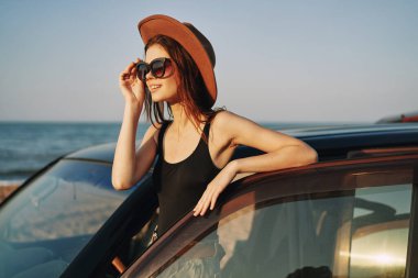 Güneş gözlüklü neşeli bir kadın ve arabanın yanındaki okyanusta bir şapka.