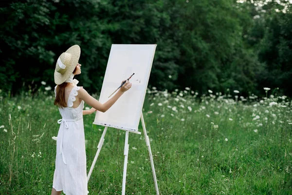 Una mujer en un vestido blanco en un campo con flores pinta un cuadro —  Fotos de Stock