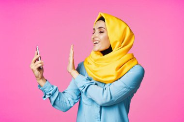 Sarı tesettürlü neşeli bir kadın. Elinde telefon, iletişim teknolojisi var.