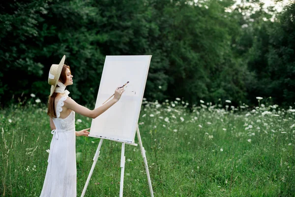 Una mujer en un vestido blanco en un campo con flores pinta un cuadro —  Fotos de Stock