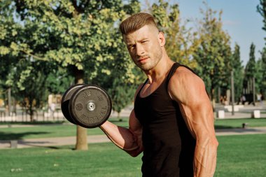 Siyah tişörtlü atletik bir adam parkta dambıllarla antrenman yapıyor.