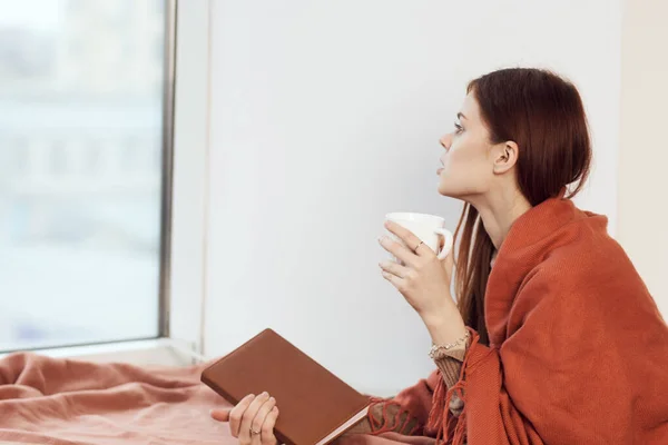 Mujer con una taza de bebida en el alféizar de la ventana leyendo un libro de descanso por la mañana — Foto de Stock
