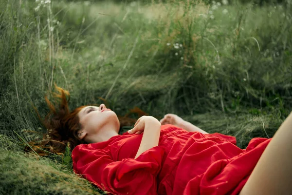 Hezká žena v červených šatech leží na trávě na poli příroda čerstvý vzduch — Stock fotografie